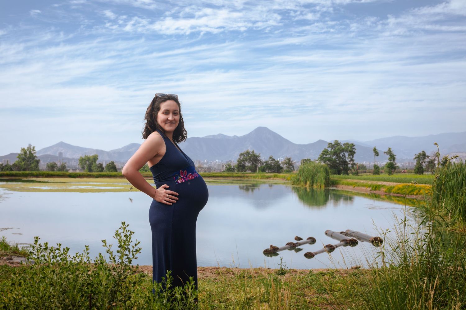 Smiling pregnant woman in navy dress posing by a scenic lake with mountains.