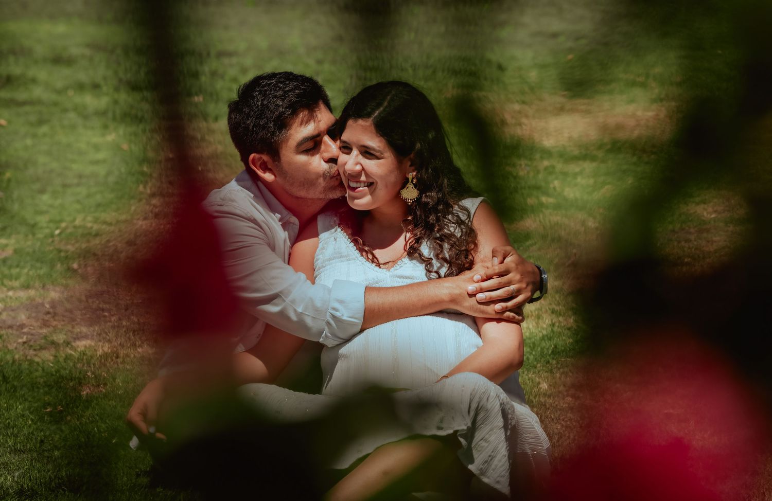 Couple sitting on grass, man kissing pregnant woman's cheek, framed by red flowers in lush green park.