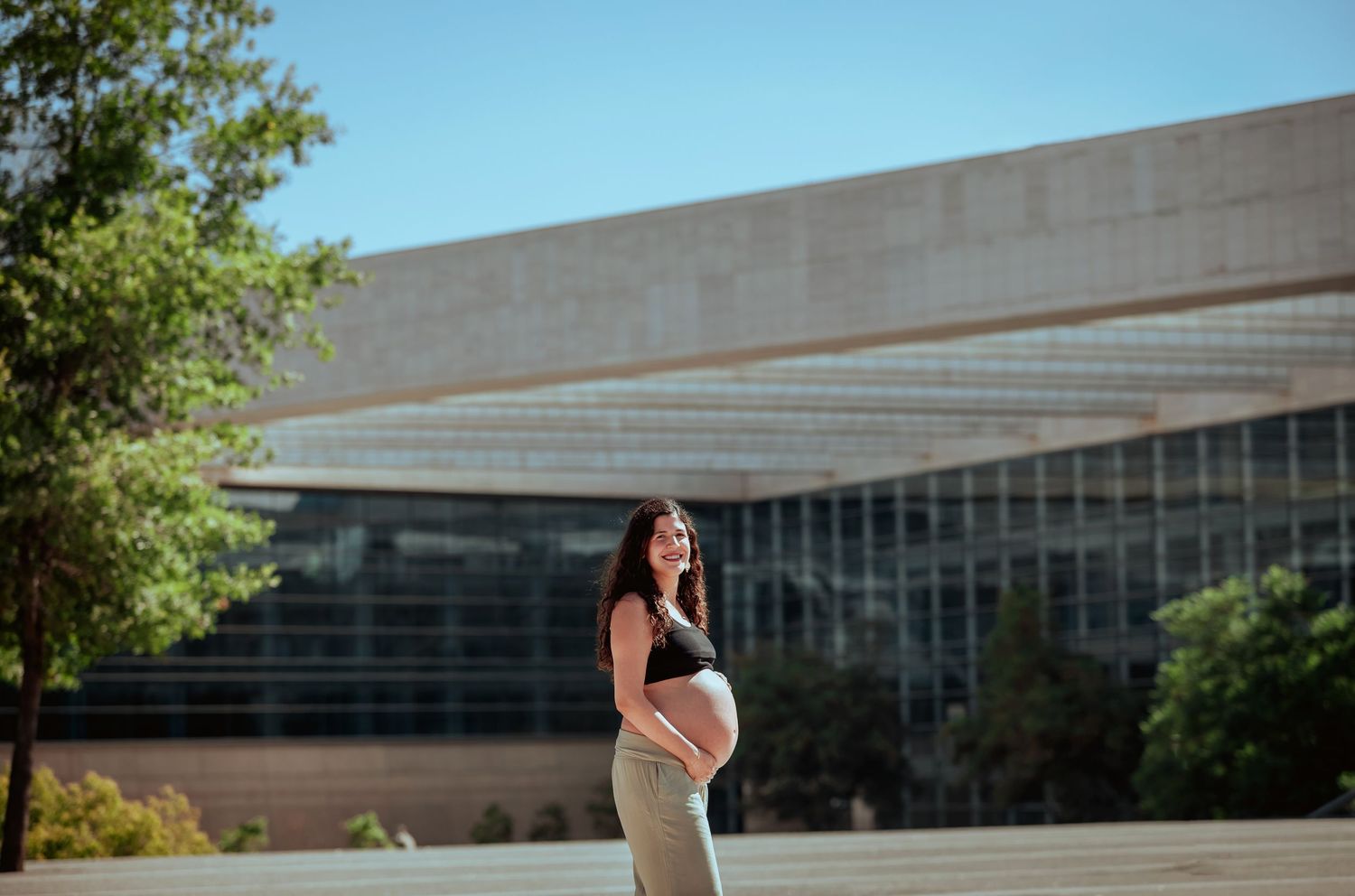 Smiling pregnant woman in black crop top and sage pants standing before a modern glass building.