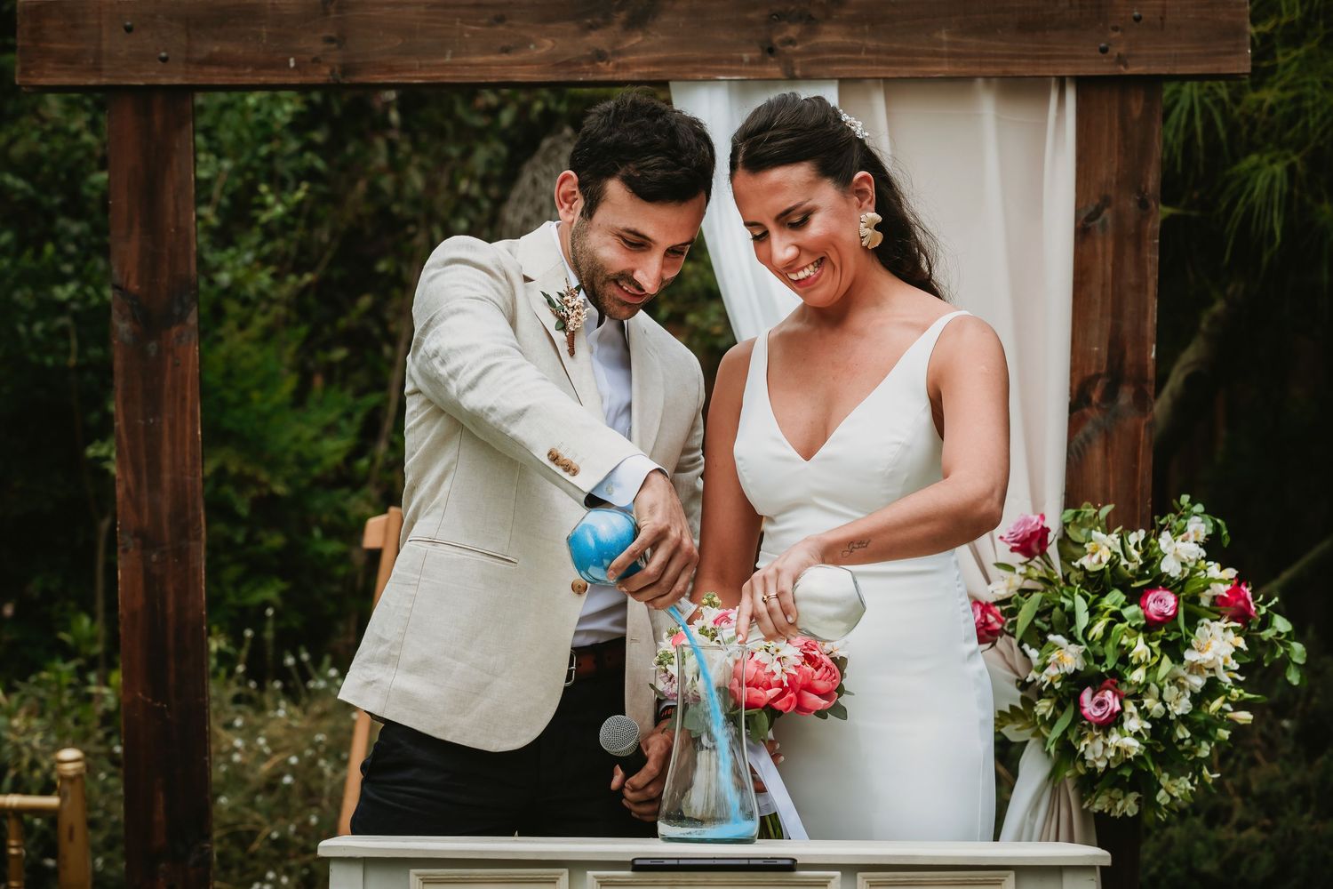 Bride and groom performing a sand ceremony at an outdoor wedding, smiling joyfully under a floral arch.