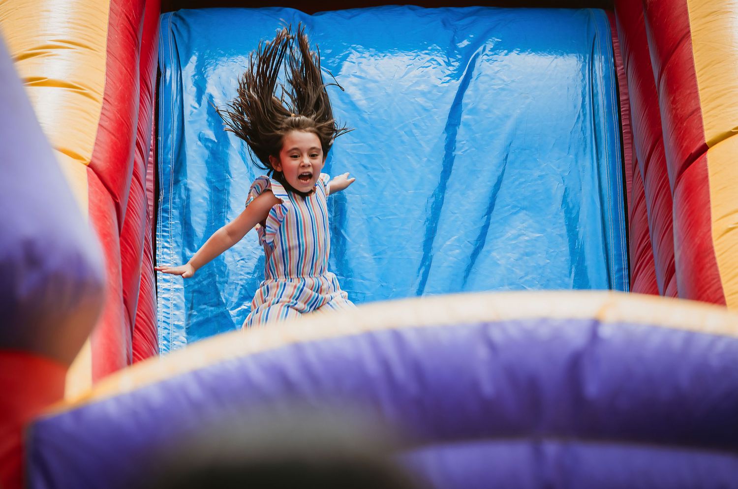 Excited girl with flying hair sliding down colorful inflatable bounce house slide at outdoor event.