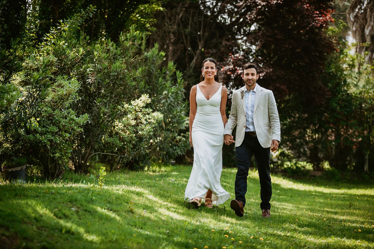 Bride and groom walking hand in hand through lush green garden on their wedding day.