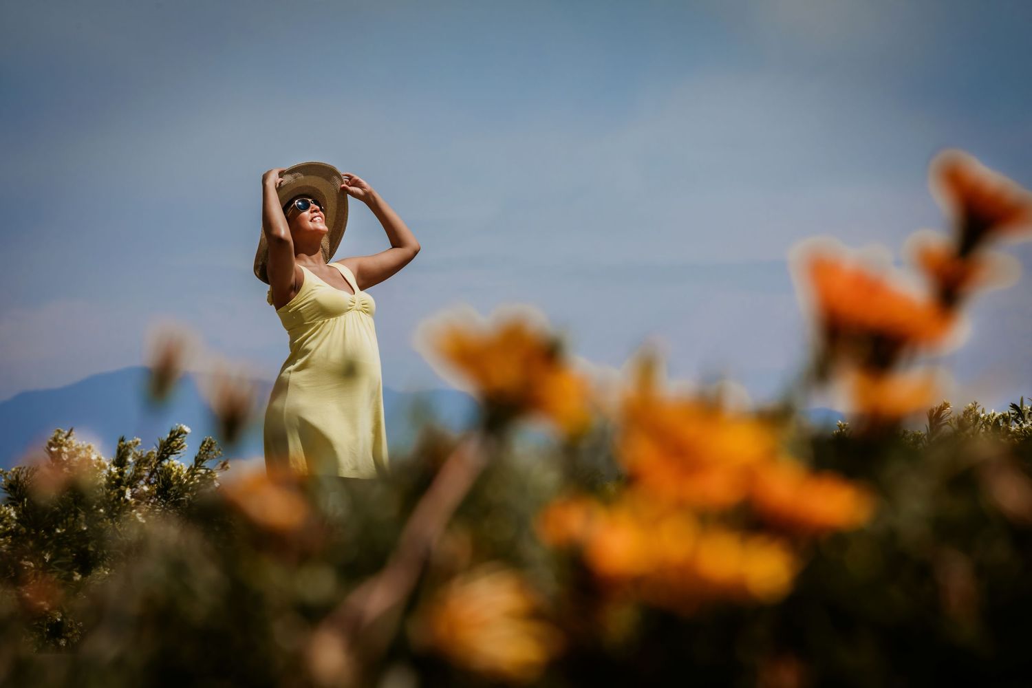 Pregnant woman in yellow dress and hat standing among vibrant orange wildflowers with mountains in the background.