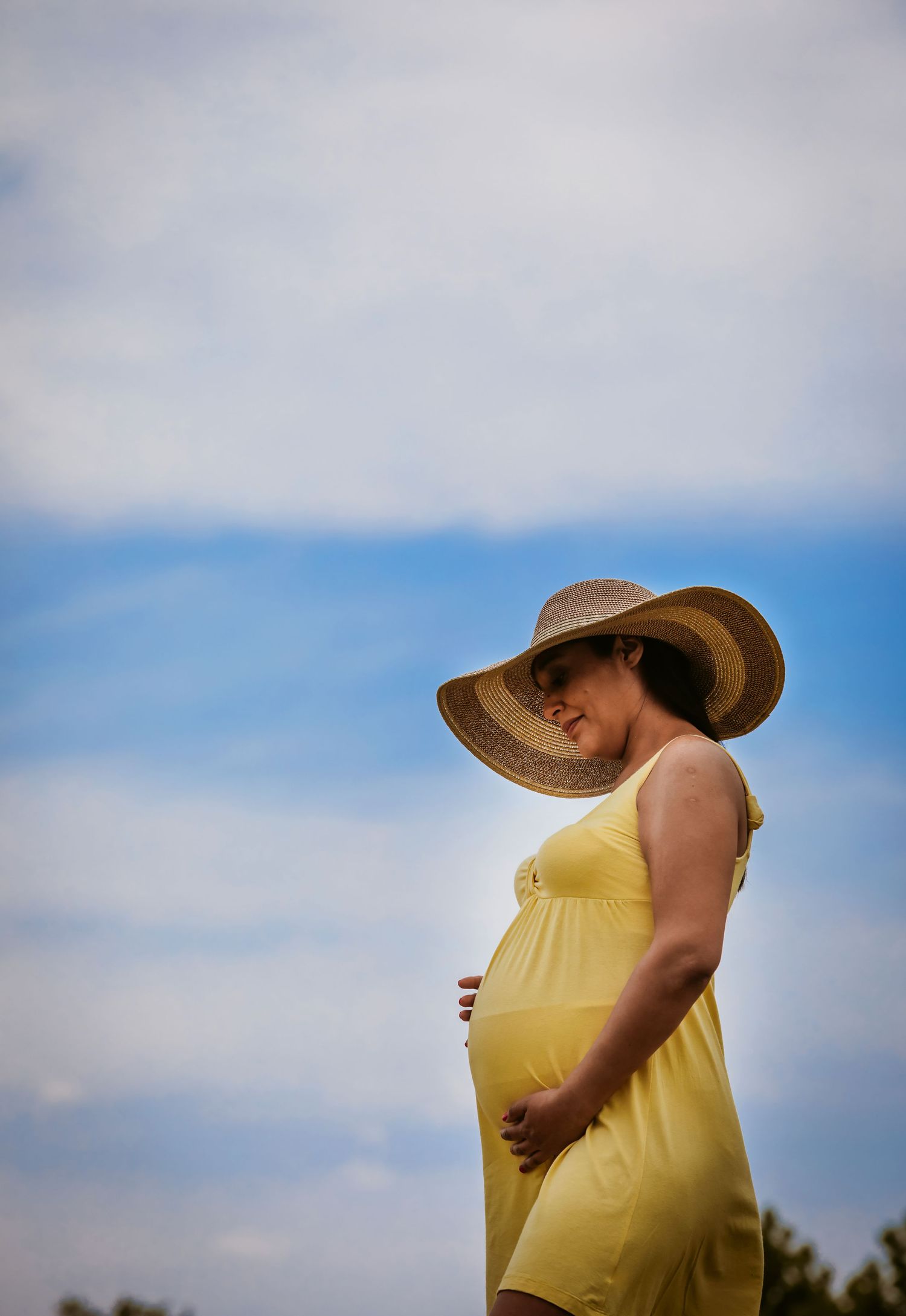 Pregnant woman in yellow dress and wide-brim straw hat cradling her baby bump against a blue sky.