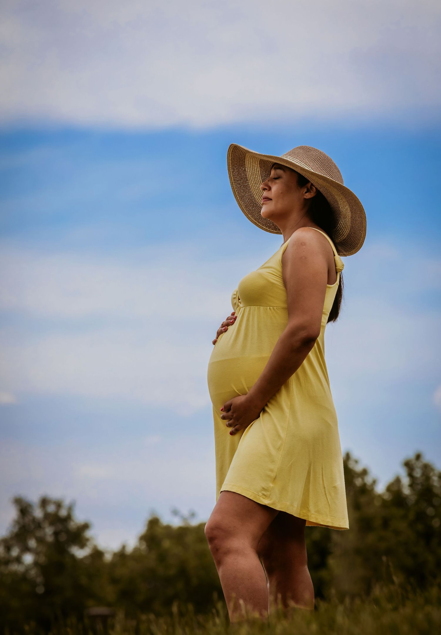 Pregnant woman in yellow dress and wide-brim hat cradling baby bump outdoors against blue sky.