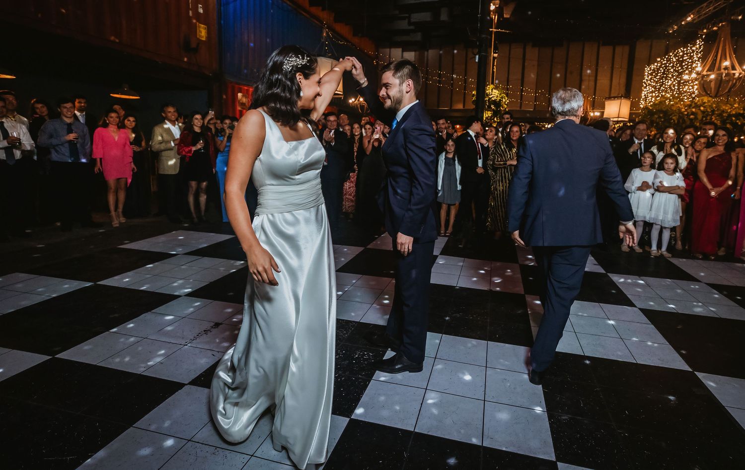 Bride in silver gown and groom in navy suit share first dance on checkered floor at elegant wedding reception.