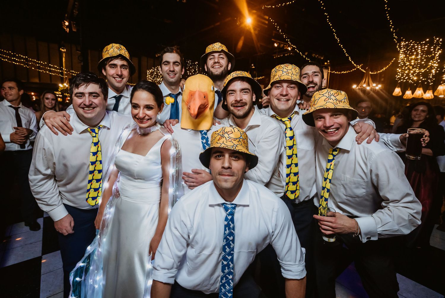 Wedding party posing with fun hats and accessories on a dance floor with string lights at a festive reception.