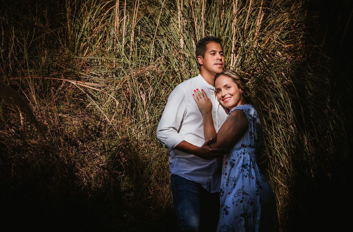 Couple embracing romantically in tall golden grass at night, woman in blue dress leaning on man in white shirt.