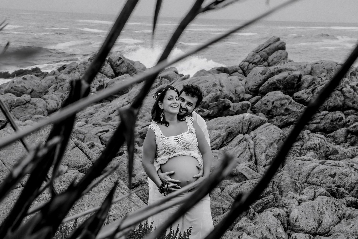 Black and white maternity photo of a couple embracing on rocky coastline with ocean waves in the background.