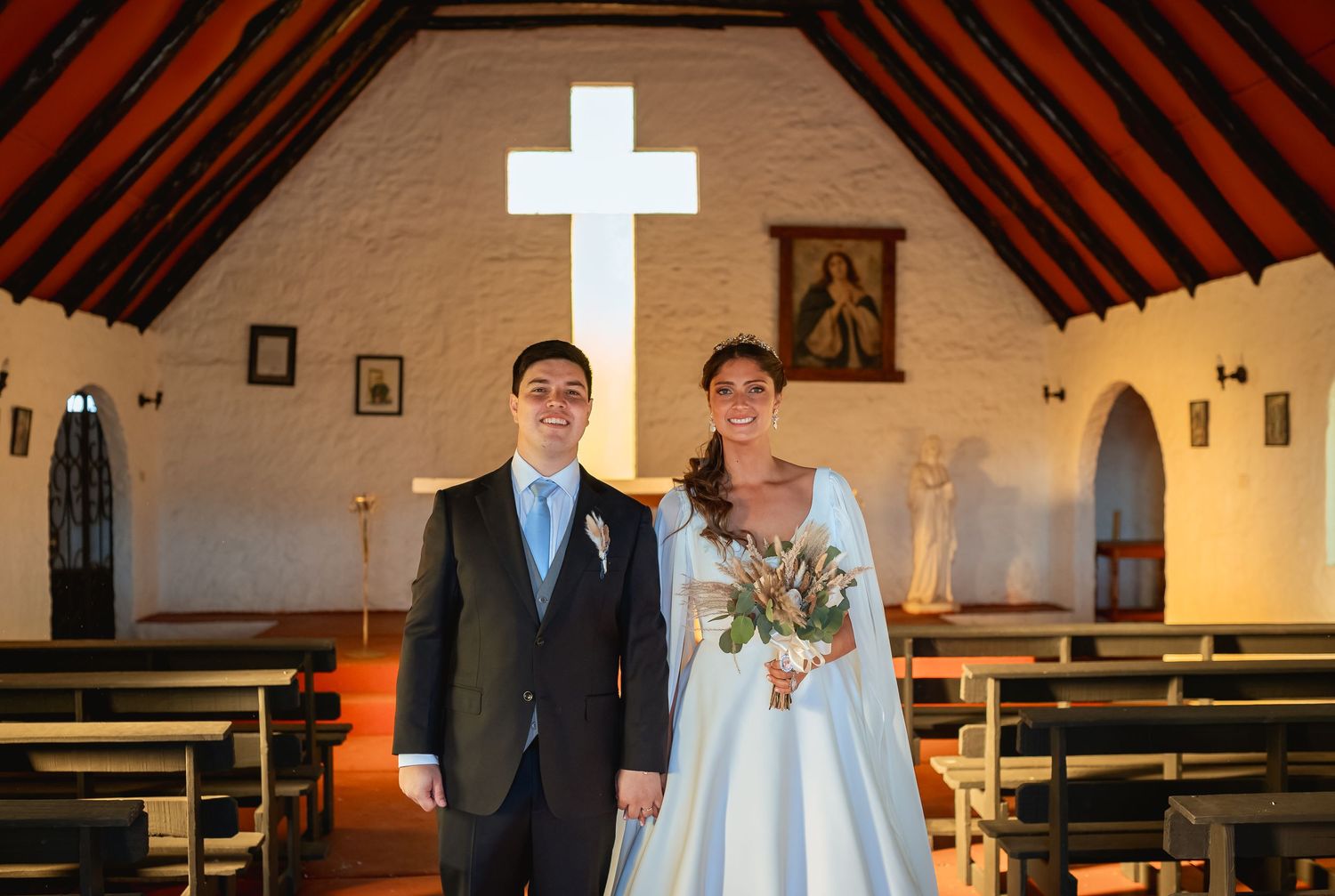 Bride and groom posing together inside a white chapel with wooden pews and an illuminated cross at the altar.