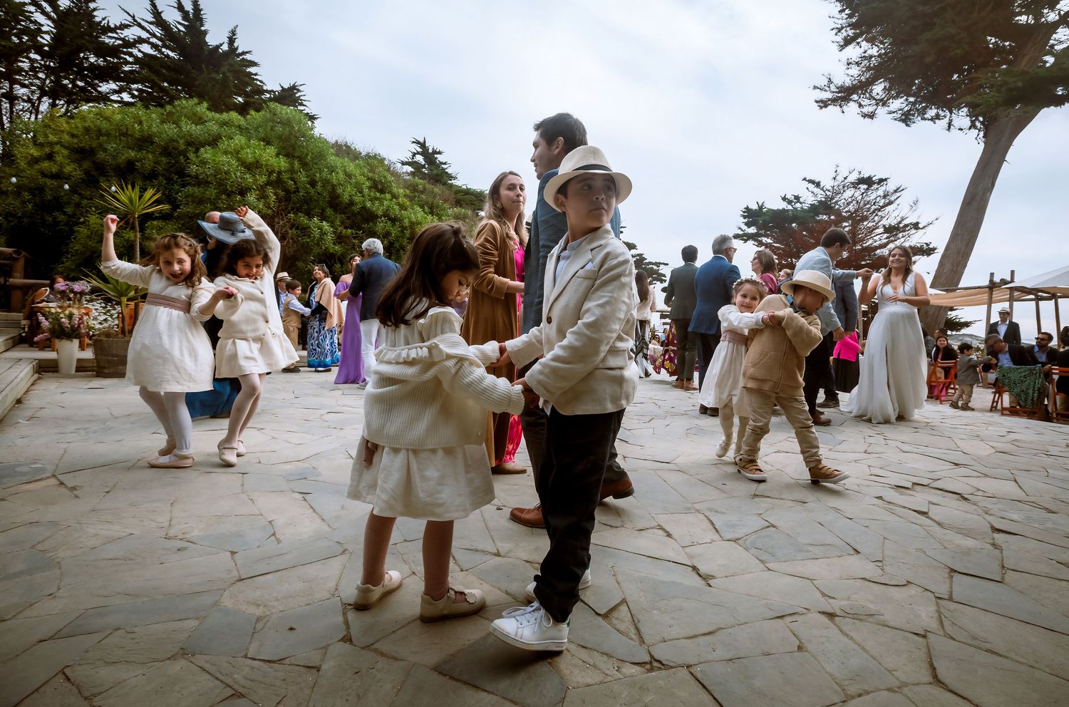 Children in formal attire dancing and playing together on a stone patio at an outdoor wedding celebration.
