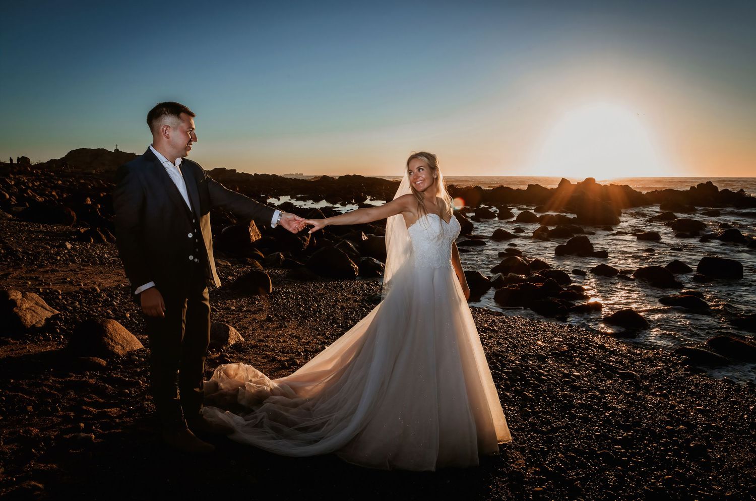 Groom leading bride by hand on rocky coastline at sunset, veil trailing behind her.