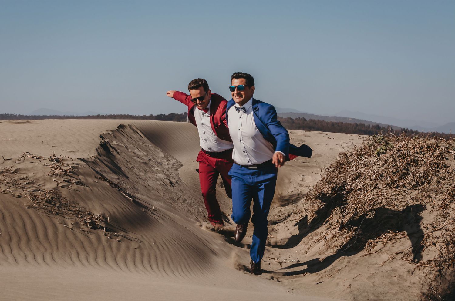 Two grooms in colorful suits running and laughing down sandy desert dunes on sunny day.