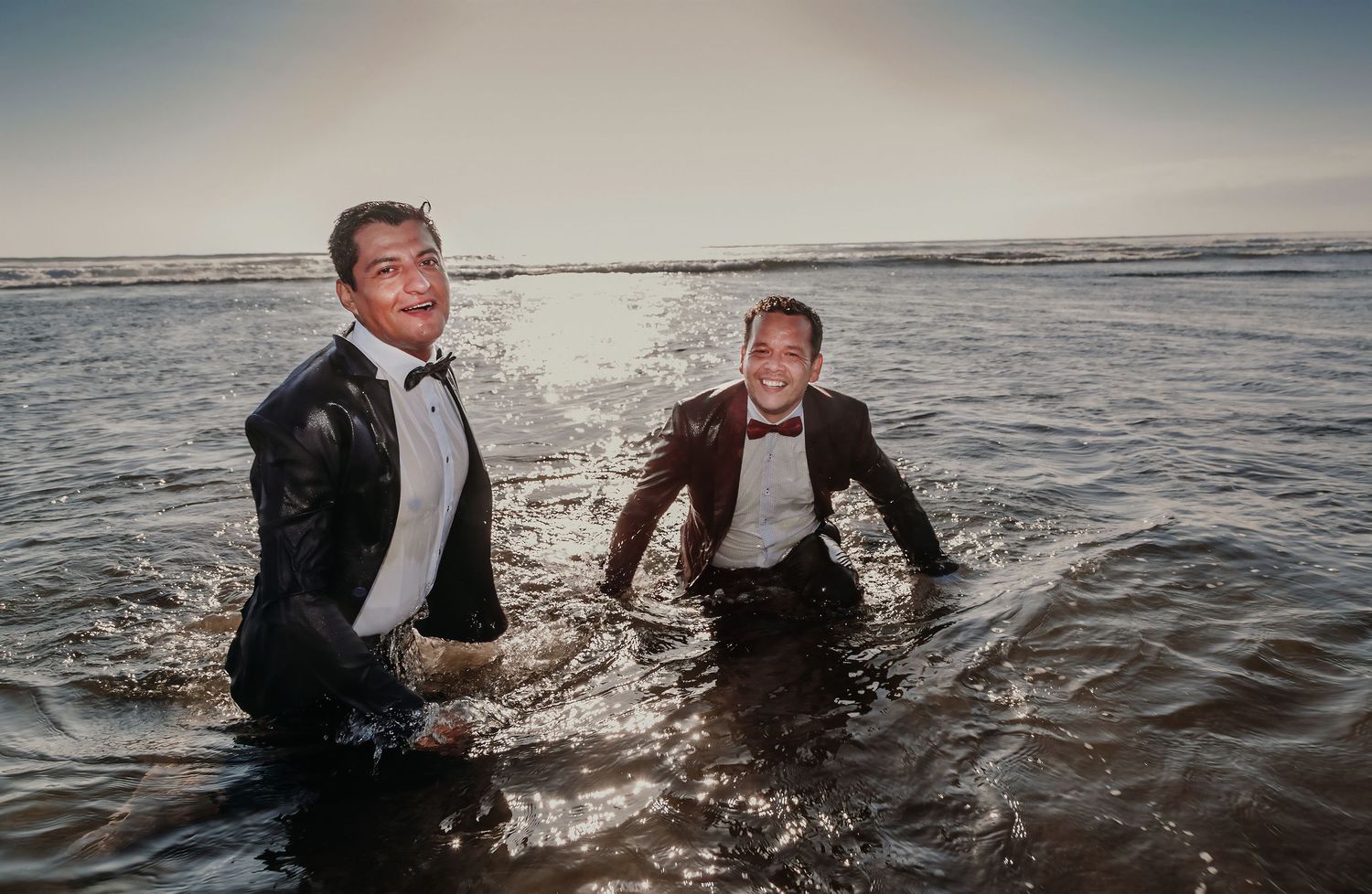 Two grooms in soaked tuxedos laugh joyfully while standing waist-deep in ocean waves.