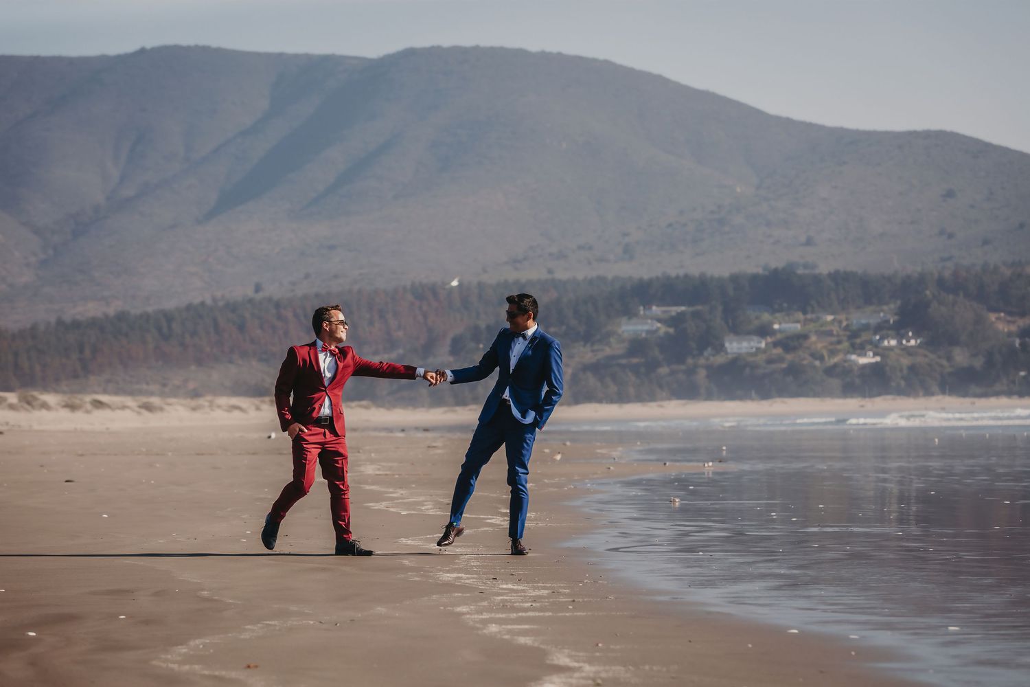 Two grooms in red and blue suits playfully interacting on a wide sandy beach with mountain views.