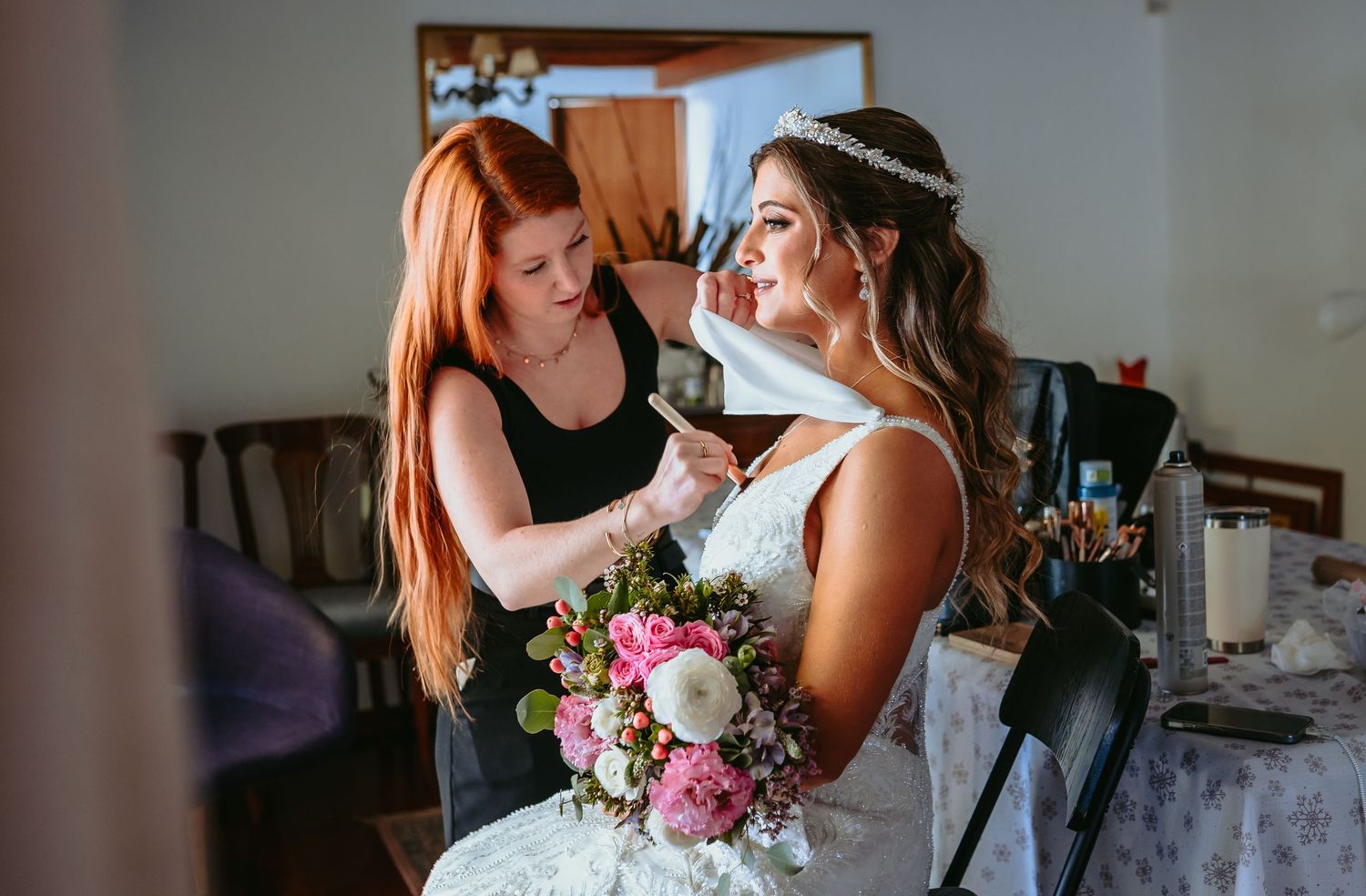 Redhead stylist doing final makeup touches on a seated bride holding a pink floral bouquet.