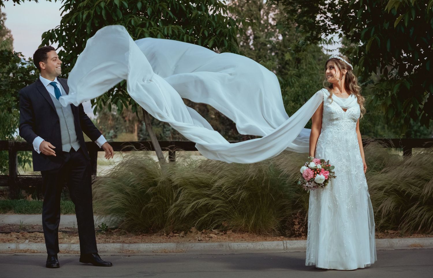 Bride's long white veil billows dramatically in the wind as couple poses on an outdoor pathway.