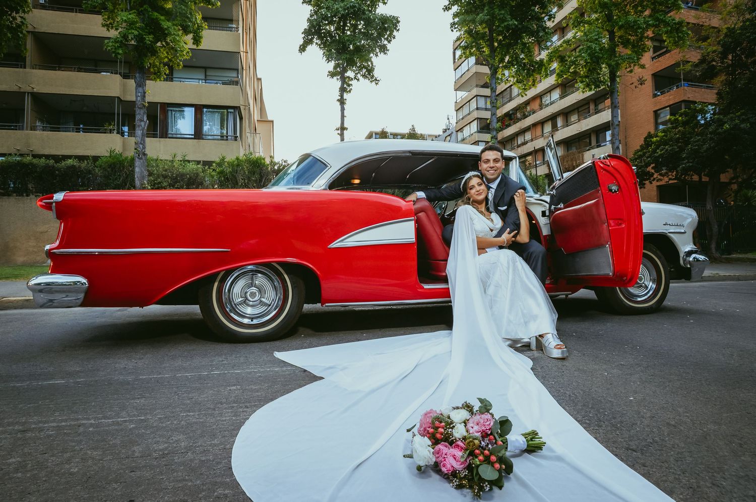 Bride and groom posing with open doors of a red vintage classic car with bridal bouquet on pavement.