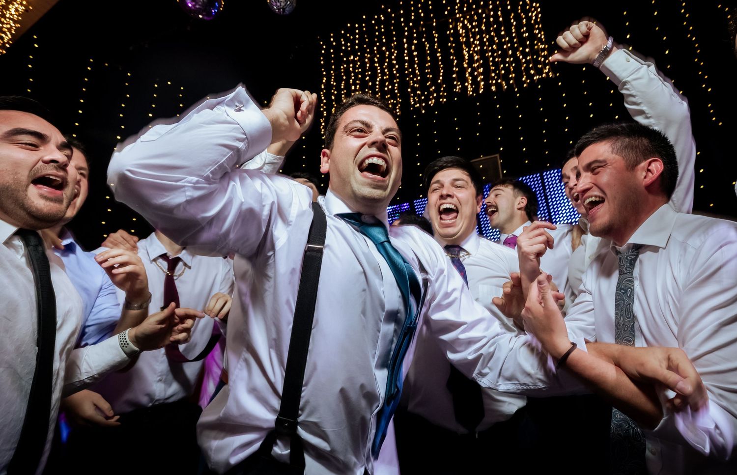 Groomsmen celebrate joyfully with raised fists and big smiles at an energetic wedding reception.