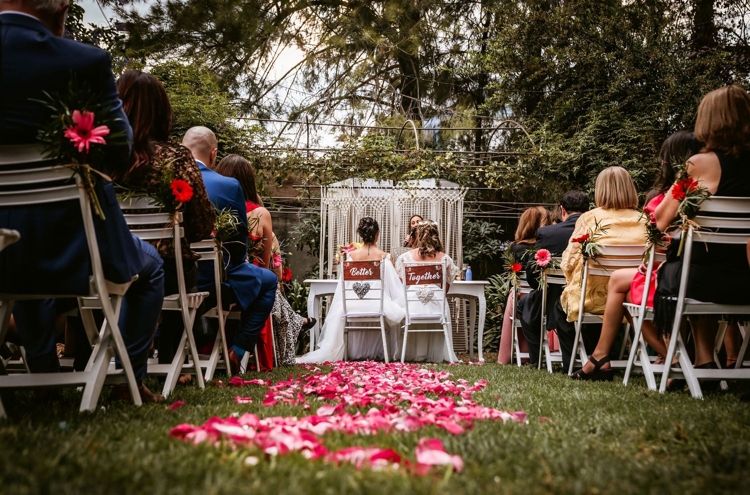 Outdoor garden wedding ceremony with guests seated in white chairs along pink rose petal aisle toward altar.