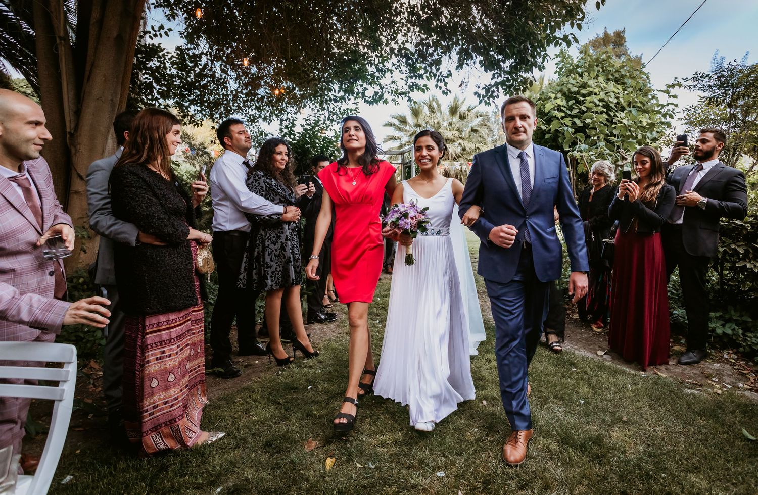 Bride walks through applauding guests with woman in red dress and man in blue suit outdoors.