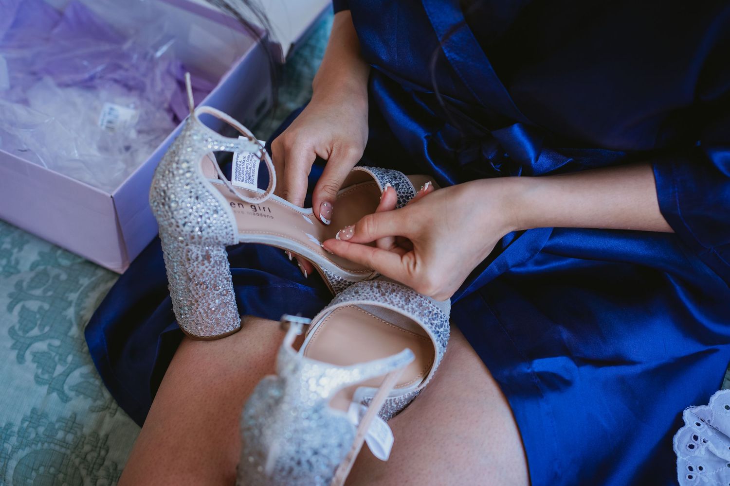 Close-up of sparkling bridal heels being fastened by a bridesmaid in a blue robe before the wedding.