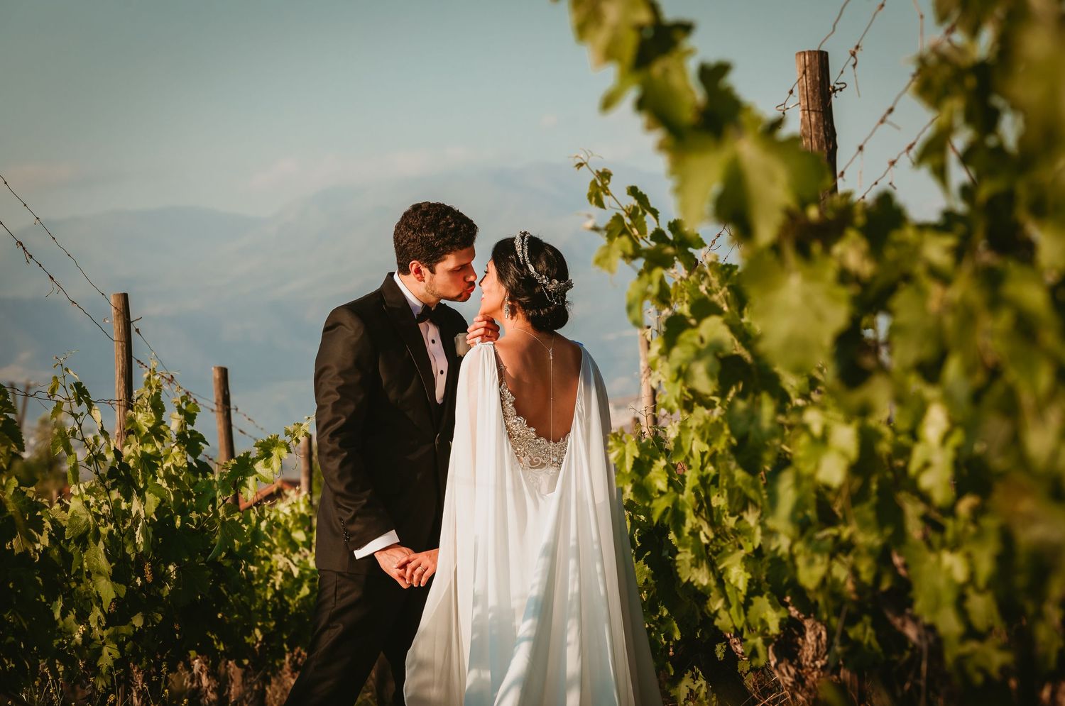 Bride and groom share a romantic almost-kiss moment surrounded by lush green vineyard vines.