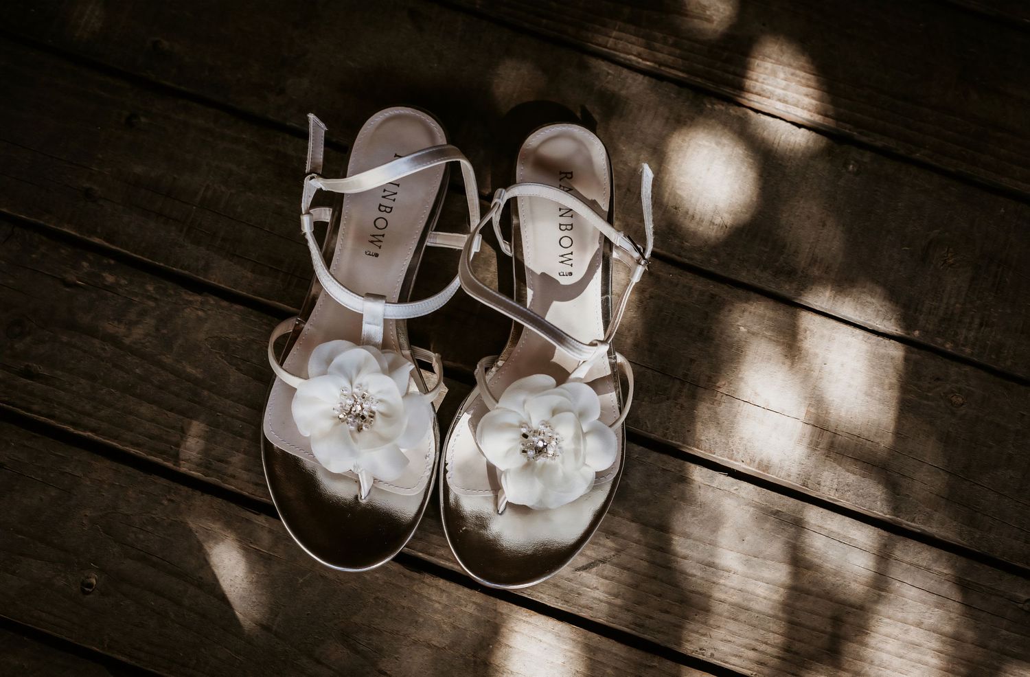 A pair of white bridal sandals with floral embellishments rest on a wooden deck in dappled sunlight.