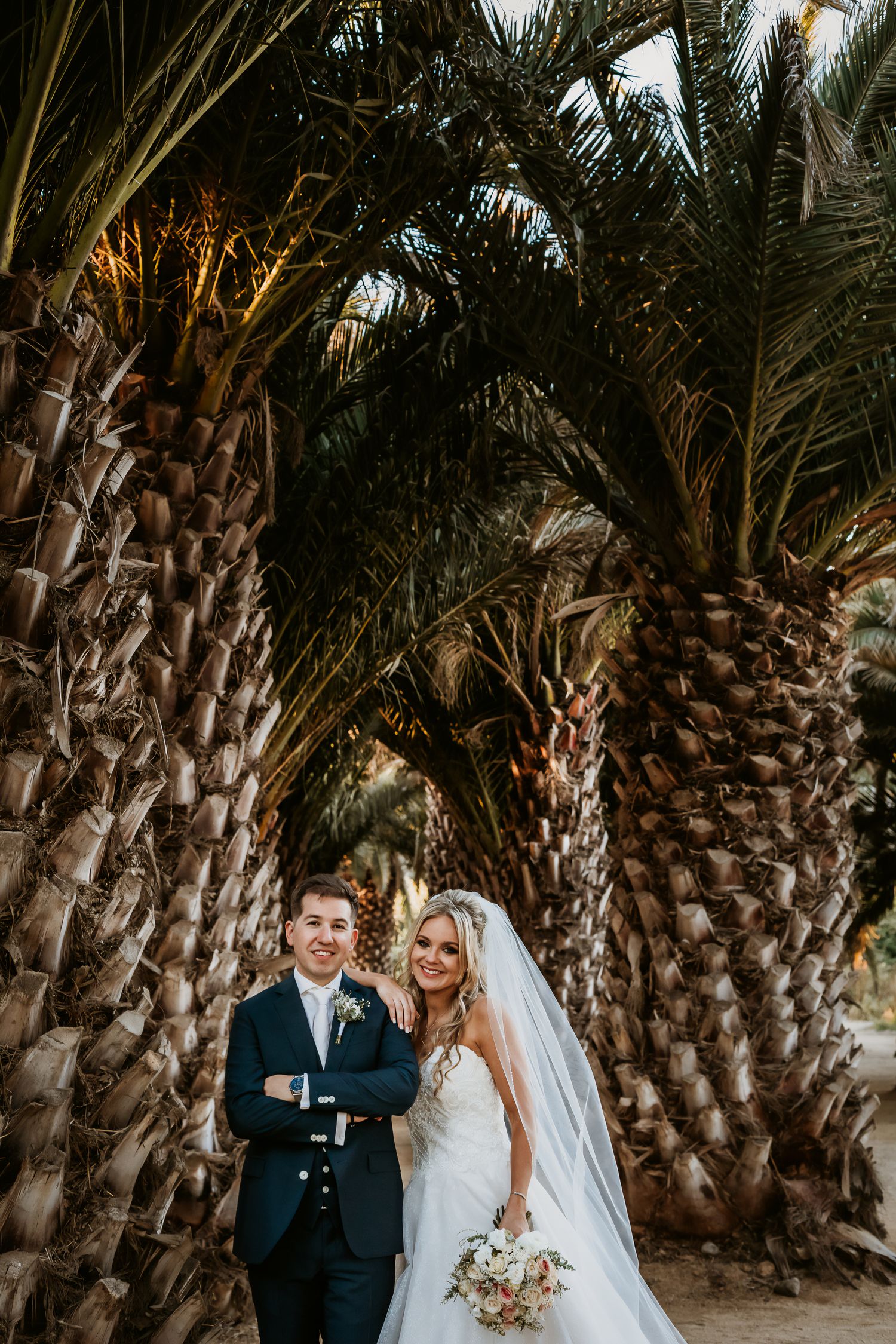 Smiling bride and groom standing together under arching palm trees with bridal bouquet.
