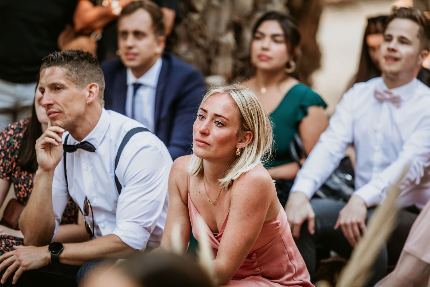 Wedding guests in formal attire listen attentively during outdoor daytime wedding ceremony.