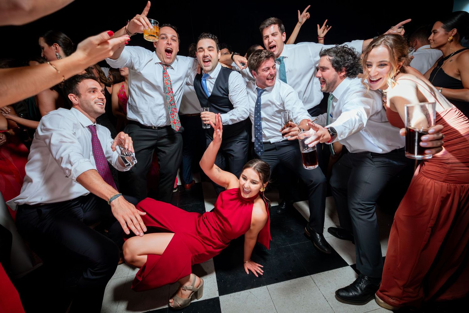 Energetic wedding guests dancing and celebrating on a checkered dance floor with a woman in a red dress.