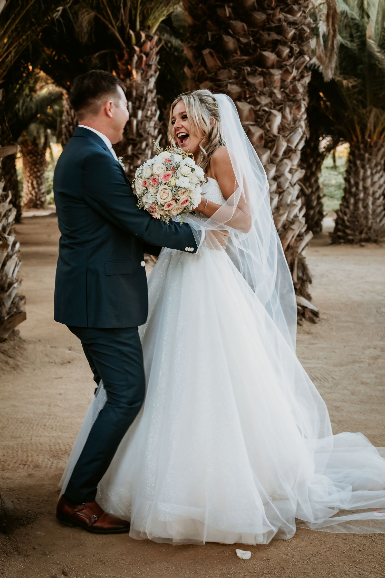 Joyful bride laughing with groom among palm trees, holding a pink and white floral bouquet.