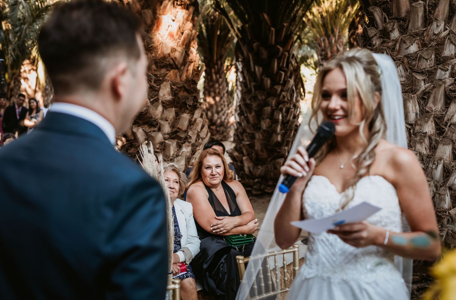 Smiling bride reads her vows into a microphone at an outdoor wedding ceremony with guests watching.