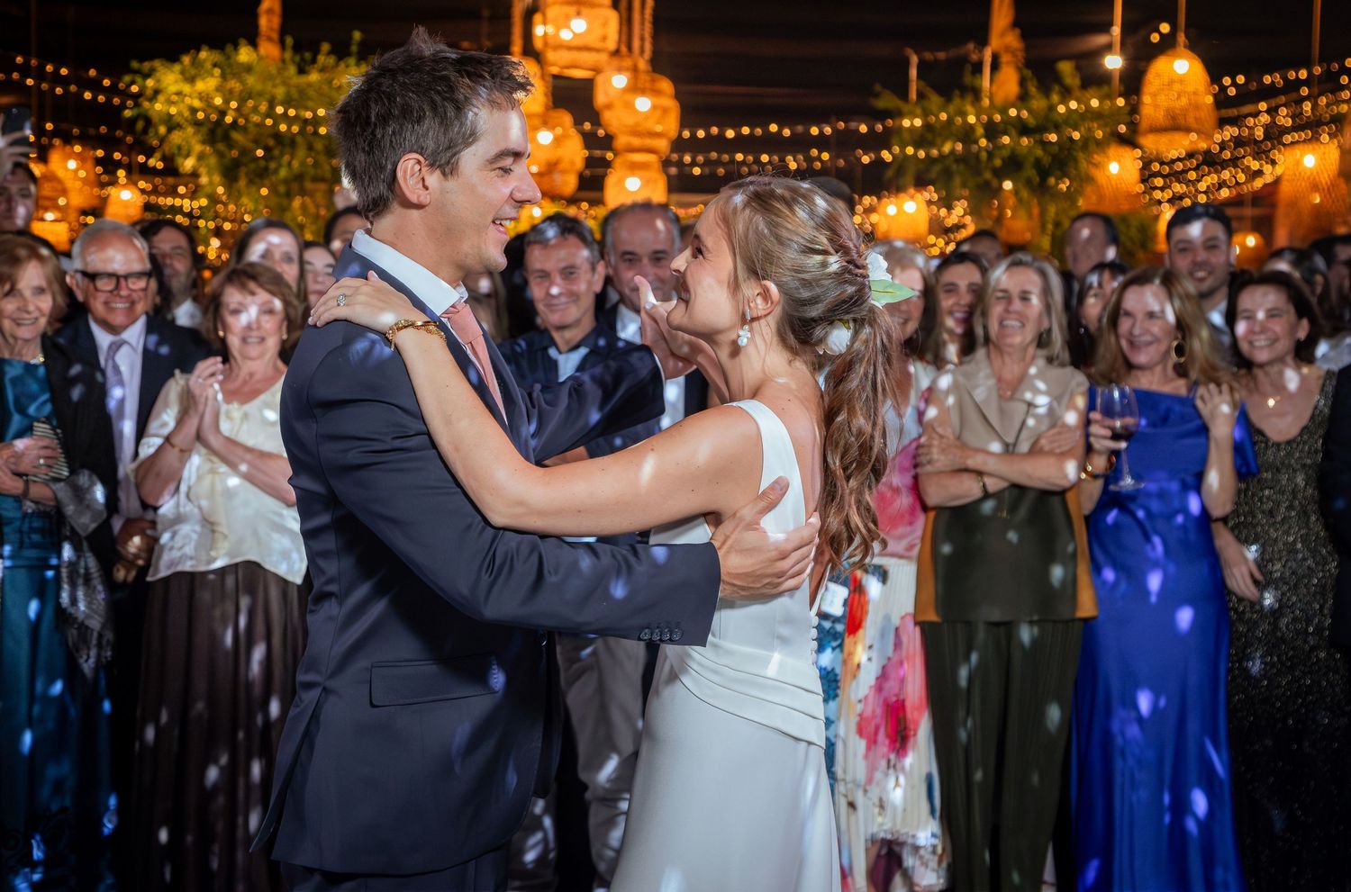 Bride and groom share their first dance surrounded by cheering guests at an outdoor wedding reception with warm lighting.