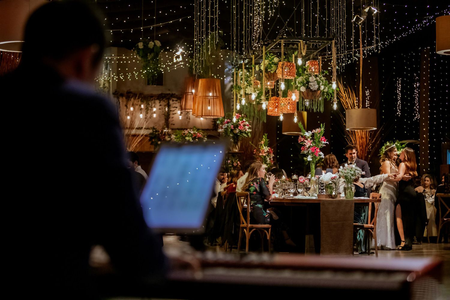 DJ oversees a beautifully decorated wedding reception hall with hanging lights and floral arrangements.