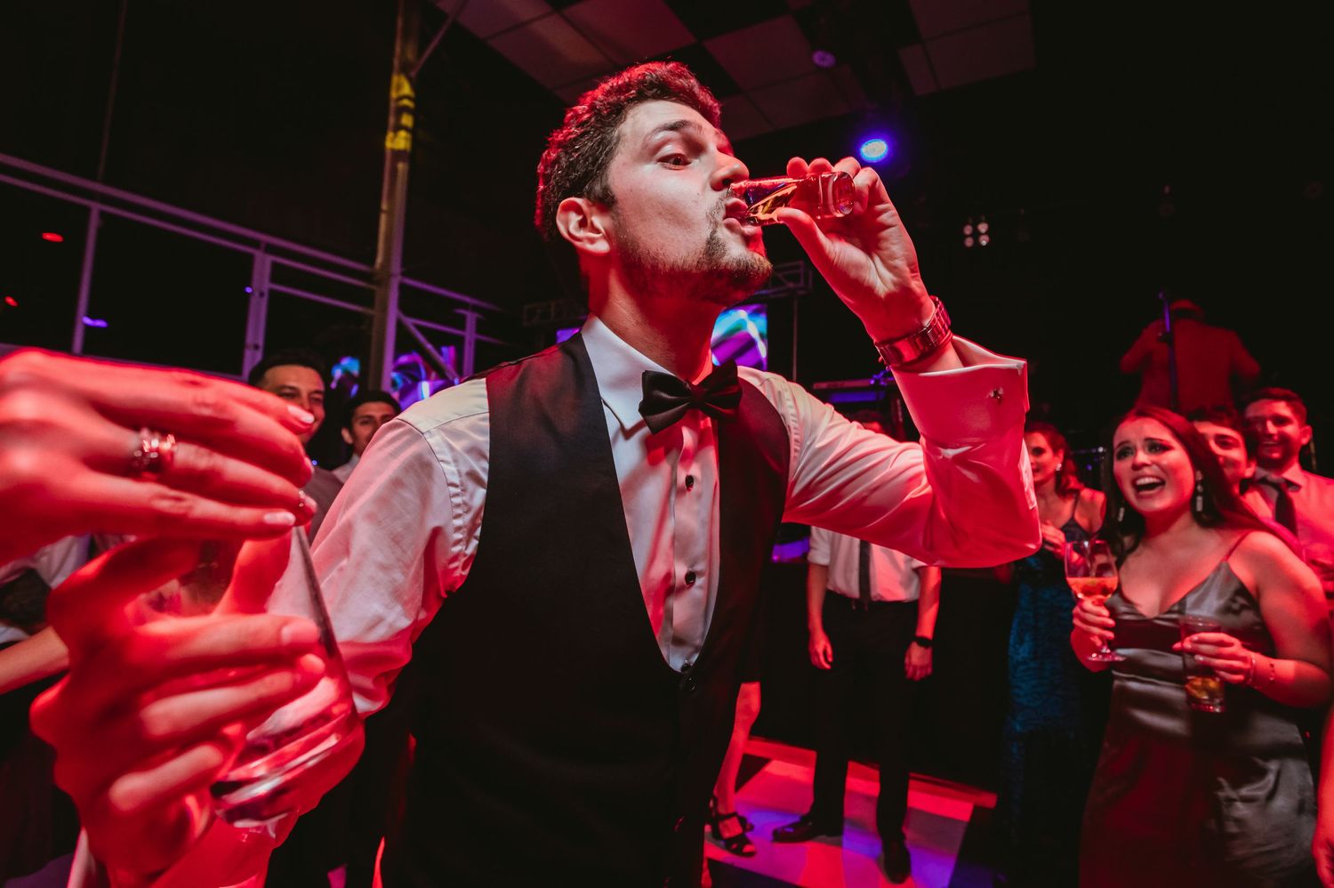 Man in tuxedo vest drinks from red cup as guests reach out at lively wedding party.
