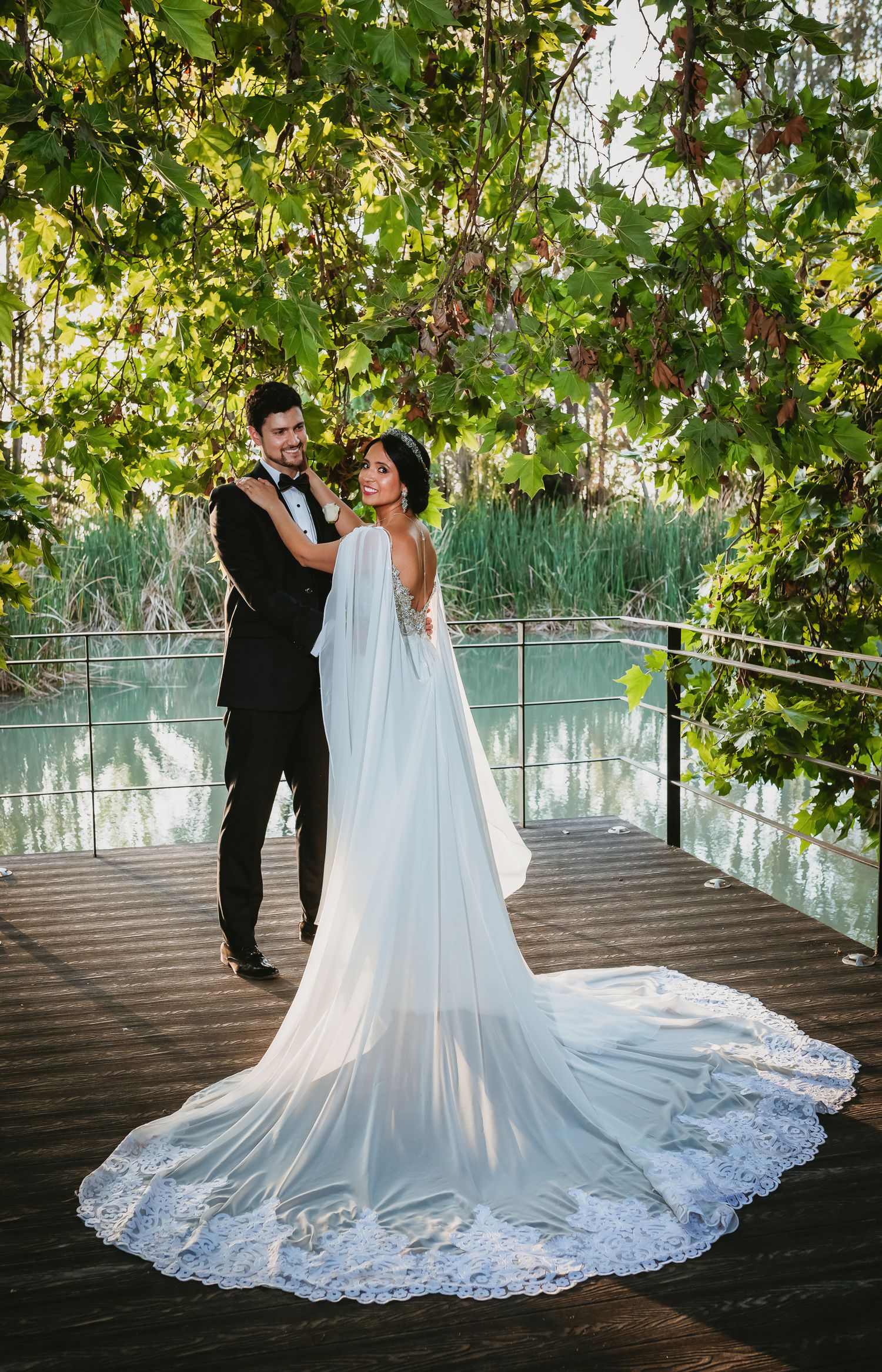 Bride in elegant white gown with lace cape train poses with groom in black tuxedo on wooden deck by serene pond.