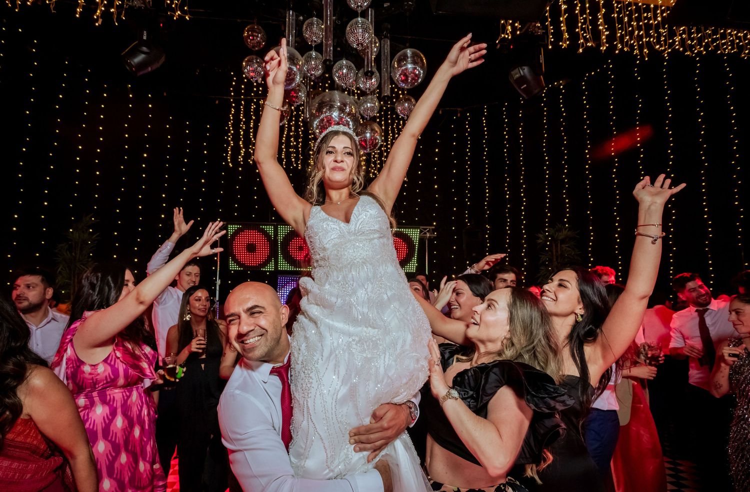 Bride lifted by guests dancing joyfully at a vibrant wedding reception with colorful lights.