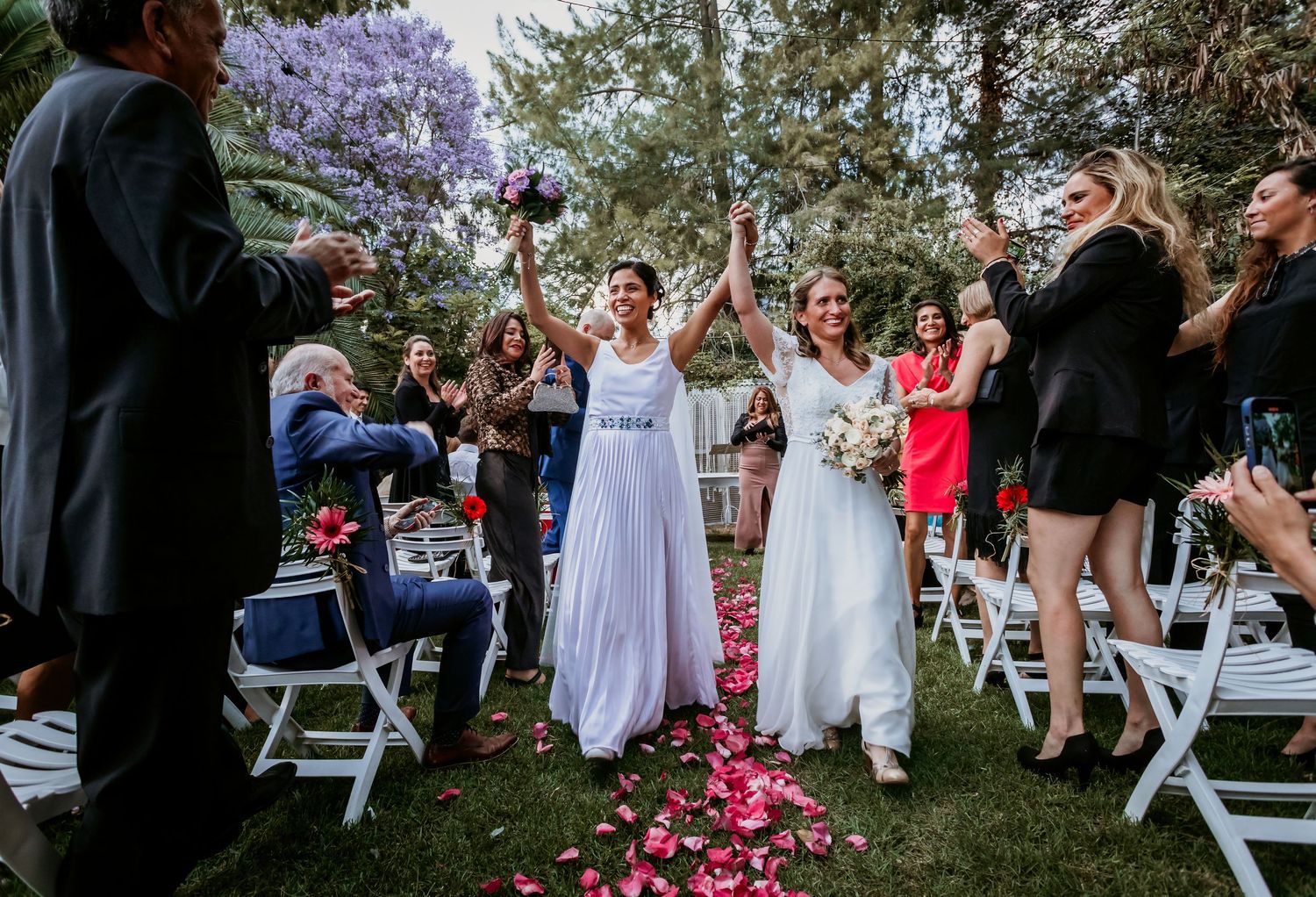 Two brides celebrating joyfully while walking down petal-strewn aisle at outdoor garden wedding ceremony.