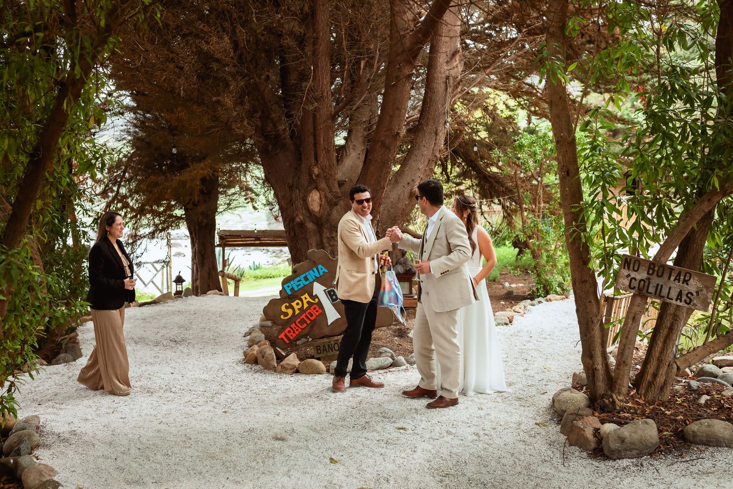 Bride and groom exchange rings in an intimate outdoor ceremony surrounded by trees and white gravel.
