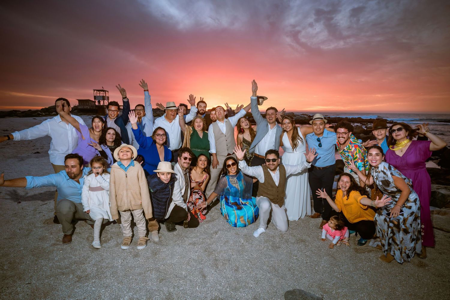 Large wedding group celebrating on beach during stunning pink and red sunset.
