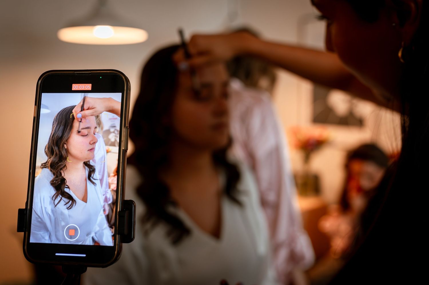 Bride getting makeup applied while being recorded on a smartphone during wedding preparations.
