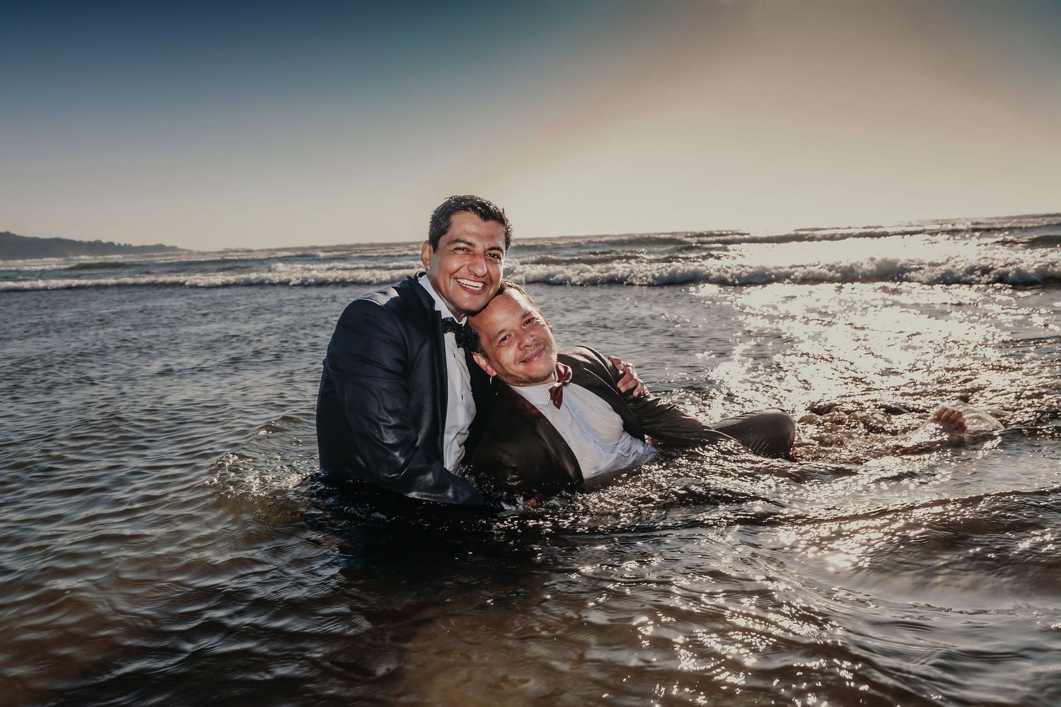 Two grooms smile warmly while sitting together in shallow ocean waves during a beach wedding shoot.
