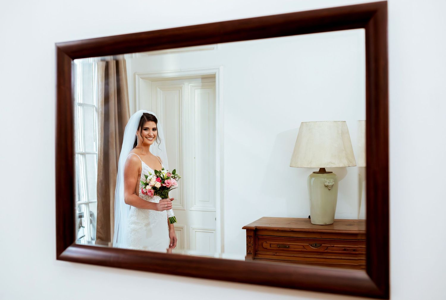 Bride in white wedding dress and veil holding bouquet reflected in ornate wooden framed mirror beside lamp.