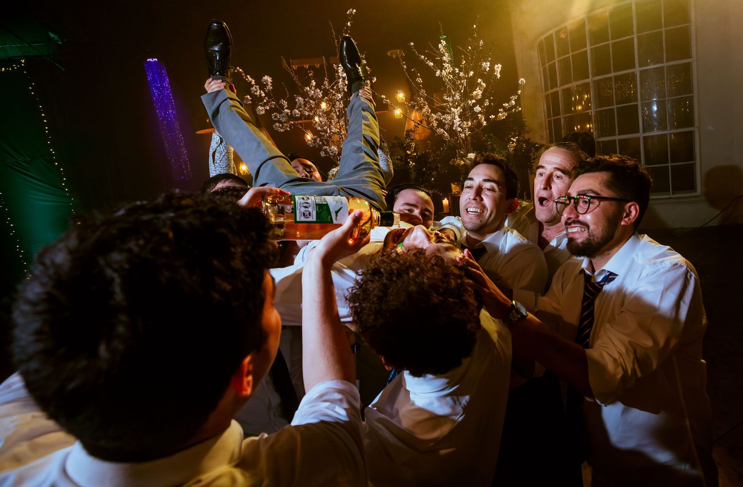 Groomsmen joyfully lifting groom at a lively outdoor wedding reception party at night.