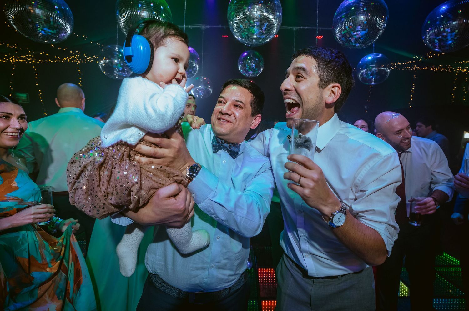 Two men celebrating at a party, one holding a baby with ear protection under disco balls.