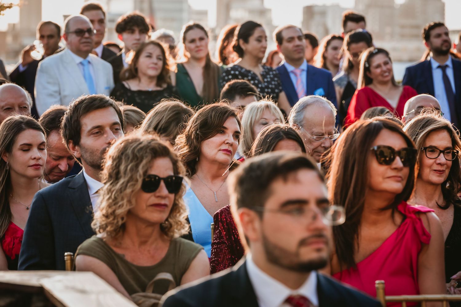 Large group of wedding guests in formal attire watching an outdoor ceremony.