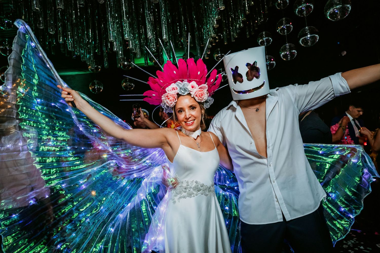 Bride in white dress with LED butterfly wings and pink floral crown poses with Marshmello-masked groom at wedding reception.