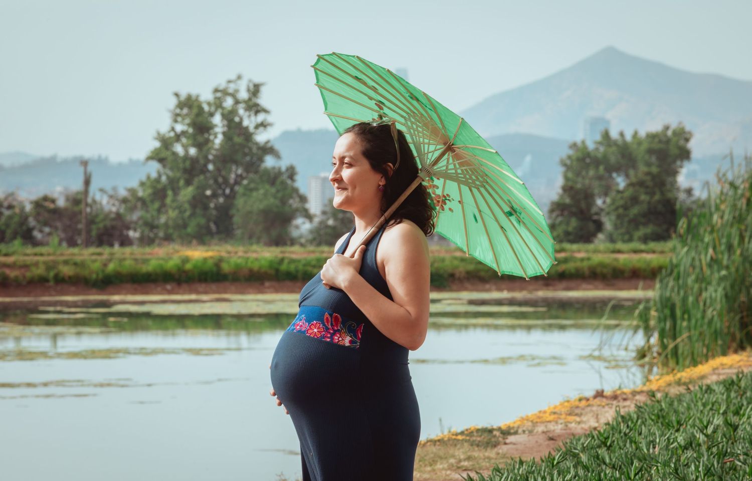 Glowing pregnant woman holding green parasol beside a calm lake with mountains.