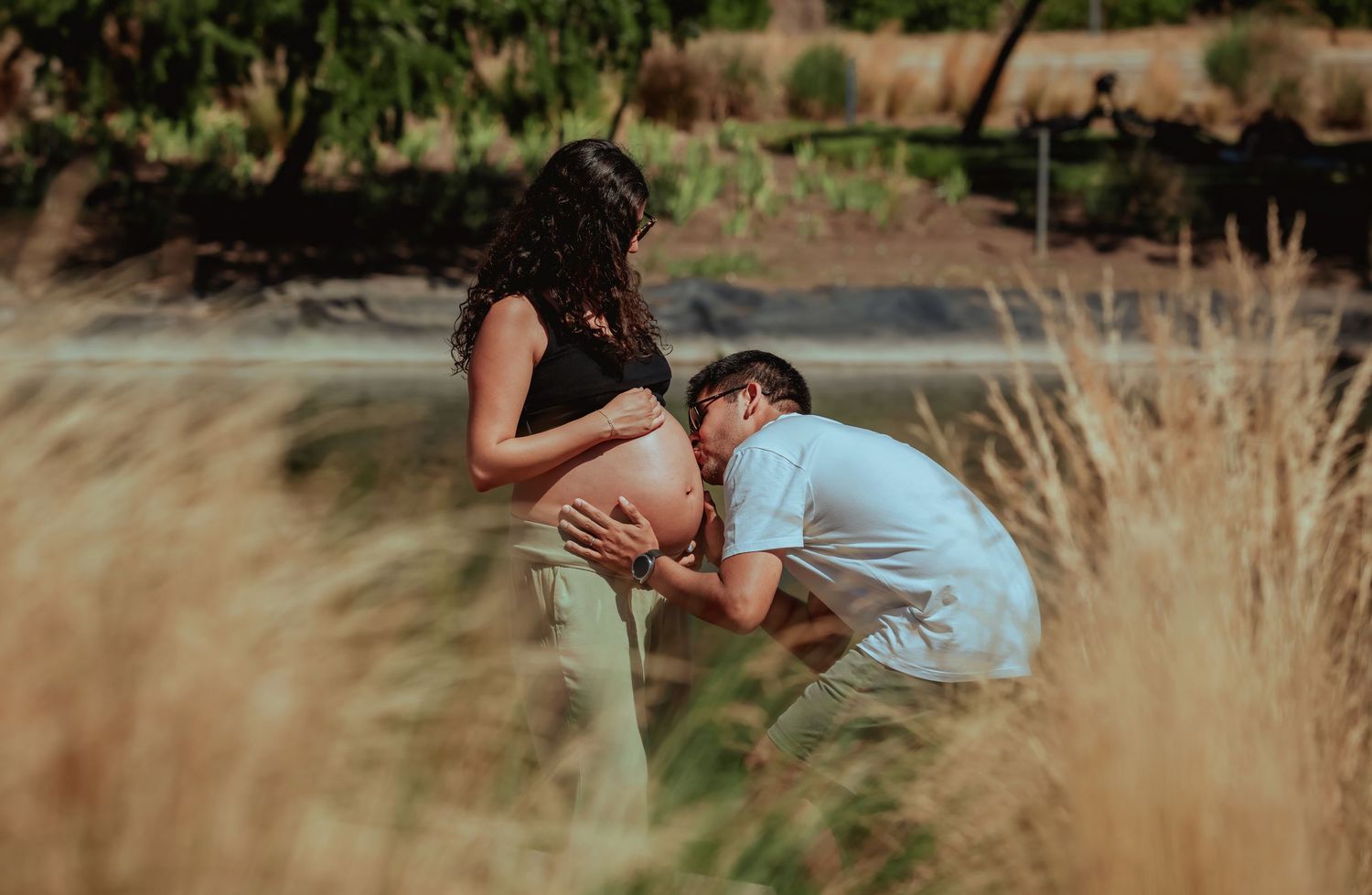 Man tenderly kissing pregnant womans belly while kneeling in a golden grass field outdoors.